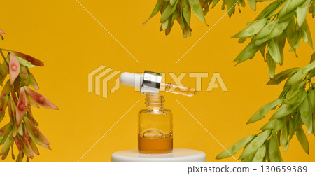 A clear glass dropper bottle with a silver cap and a white dropper dripping amber liquid, standing on a white stand. A clear glass dropper bottle with a silver cap and a white dropper dripping amber liquid, standing on a white stand. 130659389