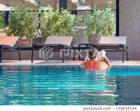 Woman in red swimsuit relaxing in indoor pool while reading book, surrounded by lounge chairs and green plants. Concept of relaxation, slow living, digital detox, self care, reading literature 130659584