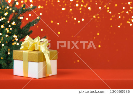 Christmas red background with green tree, white and yellow gift box with yellow satin ribbon bow and golden glitter confetti and sparkles of lights blur bokeh. Christmas and New Year holiday card 130660986