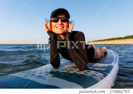 Smiling woman enjoys surfing on Baltic Sea waves, laughing joyfully lying on board with splashing water around. Bright sunlight and her laughter reflect pure joy, freedom and vibrant summer adventure 130662767