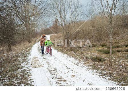 Winter adventure begins as a child learns to ride a bike along a snowy path surrounded by bare trees and a cloudy sky at an outdoor location 130662928