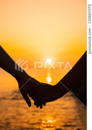 Close-up of two people holding hands against a golden sunset sky over the sea Close-up of two people holding hands against a golden sunset sky over the sea 130665978