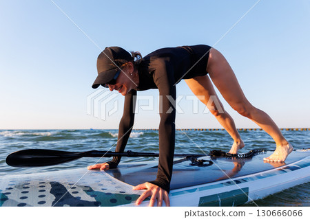 Young adult slim woman perform Yoga Downward-Facing Dog surfing SUP paddleboard Baltic Sea waves warm sunset sun lights summer day. Sunlight sea ocean waves enhance harmony body, strength mindfulness 130666066