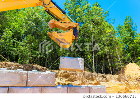 Large excavator is lifting stone wall block at construction site, set among green trees under build retaining walls. 130666713