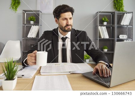 Man employee sitting with a cup of tea or coffee in office and looking at laptop monitor screen. 130666743