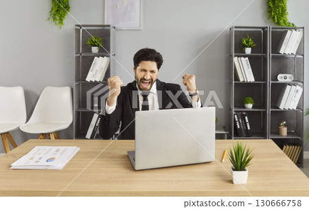 Overjoyed happy businessman clenching fists with triumph, exclaiming loudly, working on laptop 130666758
