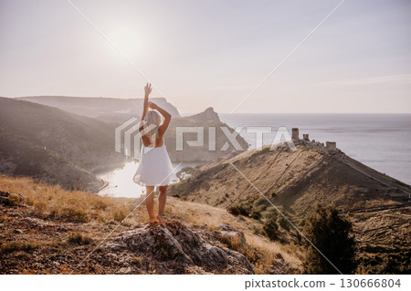 A woman stands on a hill overlooking a body of water. She is wearing a white dress and she is enjoying the view. 130666804