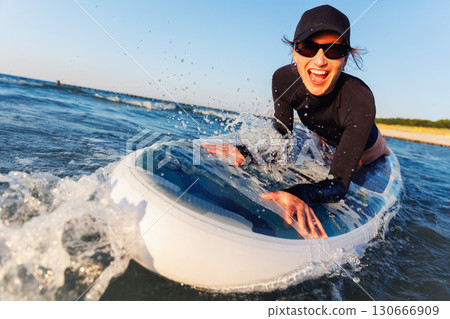 Smiling woman enjoys surfing on Baltic Sea waves, lying on board with splashing water around. Bright sunlight and her laughter reflect pure joy, freedom and vibrant summer adventure 130666909