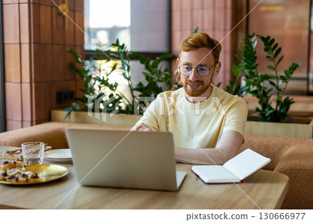 Red-haired man using laptop while eating pizza at cafe table Red-haired man using laptop while eating pizza at cafe table 130666977