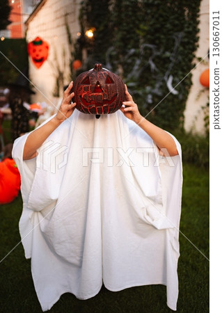Child wearing ghost costume holding jack-o'-lantern during Halloween party in the backyard Child wearing ghost costume holding jack-o'-lantern during Halloween party in the backyard 130667011