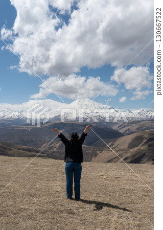 Person standing on a hill with raised arms facing Mount Elbrus on a partly cloudy day Person standing on a hill with raised arms facing Mount Elbrus on a partly cloudy day 130667522