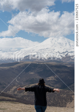 Person standing on a hill with raised arms facing Mount Elbrus on a partly cloudy day Person standing on a hill with raised arms facing Mount Elbrus on a partly cloudy day 130667525