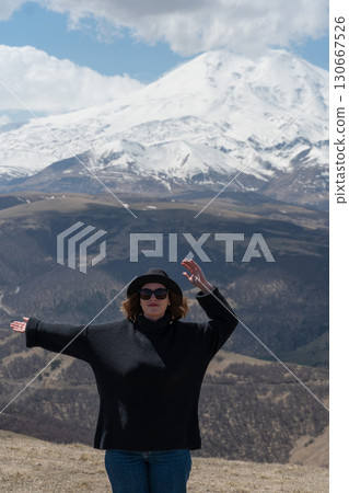 Woman in black sweater and hat enjoys panoramic view of snow-covered Mount Elbrus under a dramatic sky Woman in black sweater and hat enjoys panoramic view of snow-covered Mount Elbrus under a dramatic sky 130667526