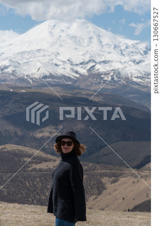 Woman in black sweater and hat enjoys panoramic view of snow-covered Mount Elbrus under a dramatic sky Woman in black sweater and hat enjoys panoramic view of snow-covered Mount Elbrus under a dramatic sky 130667527