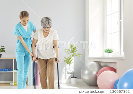 Young female caregiver helping elderly woman to walk with crutches during rehabilitation in clinic. Young female caregiver helping elderly woman to walk with crutches during rehabilitation in clinic. 130667561
