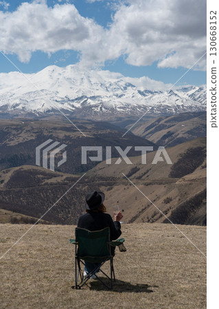 Woman in a black hat and sweater sitting in a green camping chair, enjoying a glass of white wine with a panoramic view of Mount Elbrus Woman in a black hat and sweater sitting in a green camping chair, enjoying a glass of white wine with a panoramic view of Mount Elbrus 130668152