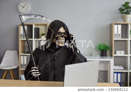 Grim reaper in black hooded cloak sitting at office desk with scythe and talking on mobile phone. Grim reaper in black hooded cloak sitting at office desk with scythe and talking on mobile phone. 130668212