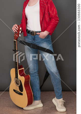 Front view of low section of female musician's body, with classical guitar. Stylish musician wearing red leather jacket and holding guitar with one hand while keeping other in pocket of her blue jeans 130668300