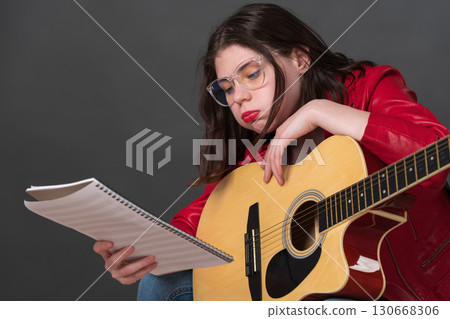 Female musician, equipped with guitar, meticulously examines sheet music while exhibiting countenance indicative of exhaustion during protracted and arduous rehearsal 130668306