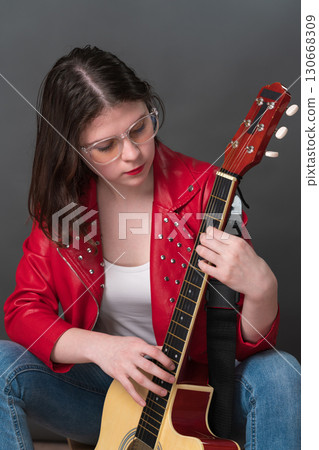 Female guitarist and musician in process of tuning her acoustic guitar, holding it upright and plucking strings in order to ascertain sound of her preferred musical instrument 130668309