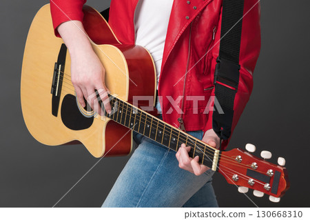 Unrecognizable woman guitarist playing acoustic guitar with concentration. Cropped view of midsection of stylish female rock musician dressed in red leather jacket and blue jeans 130668310