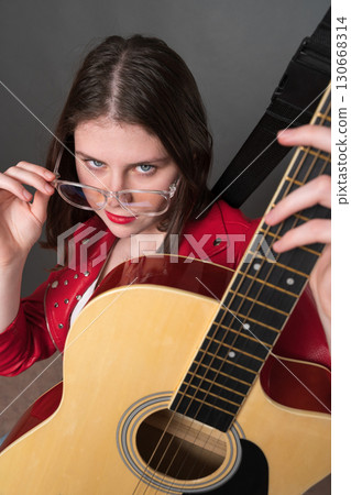 Young female guitarist holding six string classical guitar and looks at camera, lowering her glasses and holding them by temple with one hand. Confidence woman musician dressed in red leather jacket Young female guitarist holding six string classical guitar and looks at camera, lowering her glasses and holding them by temple with one hand. Confidence woman musician dressed in red leather jacket 130668314