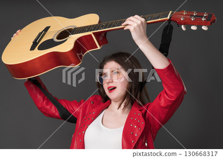 Female guitarist raised her six string acoustic guitar above her head and sings song. A female musician in glasses and a in red leather jacket Female guitarist raised her six string acoustic guitar above her head and sings song. A female musician in glasses and a in red leather jacket 130668317