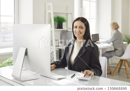Happy smiling business woman accountant working on a laptop with at office and looking at camera. 130668890