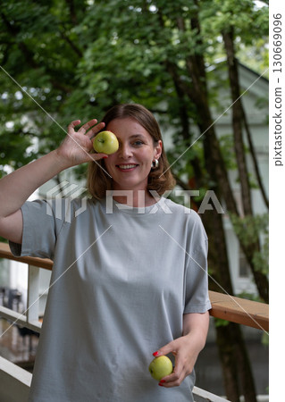 Smiling woman in a gray T-shirt playfully holds a green apple in front of one eye, while holding another in her hand, standing on a balcony 130669096