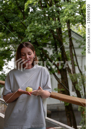 Smiling woman stands on a veranda in Peredelkino, looking down at a green apple gently held in her cupped hands 130669100