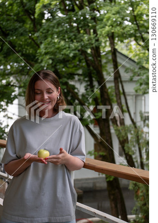 Smiling woman stands on a veranda in Peredelkino, looking down at a green apple gently held in her cupped hands 130669110