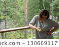 Young woman with short hair and red manicure smiles while looking at a green apple in her hand, standing on a wooden terrace surrounded by forest 130669117