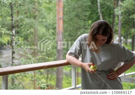 Young woman with short hair and red manicure smiles while looking at a green apple in her hand, standing on a wooden terrace surrounded by forest 130669117