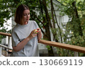 Young woman with short hair and red nails gently holds a green apple while standing on a wooden veranda, surrounded by lush greenery 130669119