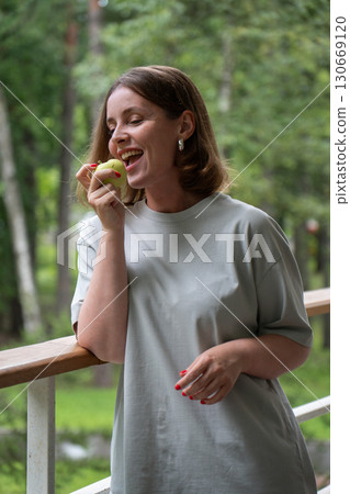 Smiling woman with short hair and silver earrings holding a green apple on a forest balcony, enjoying a moment of joy 130669120