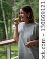 Young woman with red nails and silver earrings smiles brightly while holding a green apple on a forest balcony in Peredelkino 130669121
