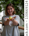 Woman gently presenting a green apple in her cupped hands, smiling with greenery in the background 130669124