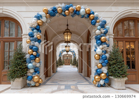 Elegant holiday arch adorned with blue, gold, and white decorations at the entrance of a luxurious building, framed by lush evergreen trees and vintage lanterns 130669974