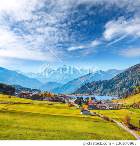 Fabulous autumn view of St.Valentin village and Haidersee (Lago della Muta) lake with Ortler peak on background. 130670965