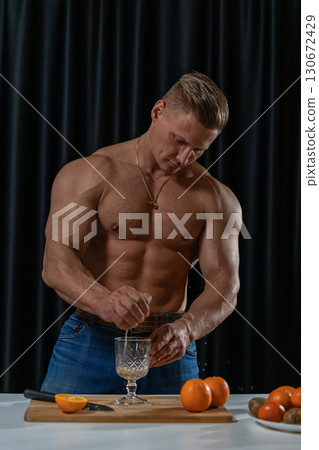 A muscular man in jeans squeezes an orange into a glass while standing in a kitchen. Fresh oranges are visible on the table showcasing a vibrant cooking scene. A muscular man in jeans squeezes an orange into a glass while standing in a kitchen. Fresh oranges are visible on the table showcasing a vibrant cooking scene. 130672429