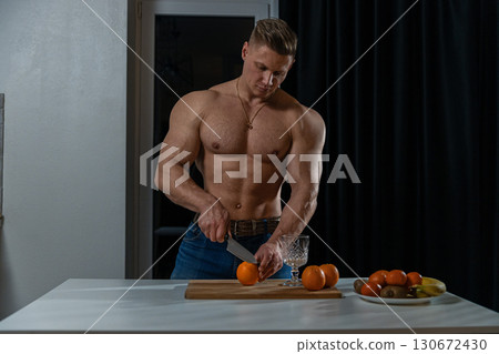 In a contemporary kitchen setting a muscular man slices an orange on a wooden cutting board. The dim evening light highlights his defined physique as he focuses on preparing fresh fruit. In a contemporary kitchen setting a muscular man slices an orange on a wooden cutting board. The dim evening light highlights his defined physique as he focuses on preparing fresh fruit. 130672430