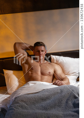 man with a bare chest is lounging in bed surrounded by pillows. The soft morning light filters in creating a calm atmosphere for his relaxed demeanor. man with a bare chest is lounging in bed surrounded by pillows. The soft morning light filters in creating a calm atmosphere for his relaxed demeanor. 130672438