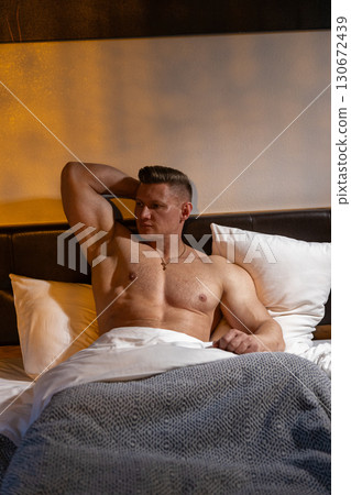 man with a muscular build is lounging in bed with his bare chest visible. He leans back against the headboard surrounded by soft pillows and blankets as morning light filters in. 130672439