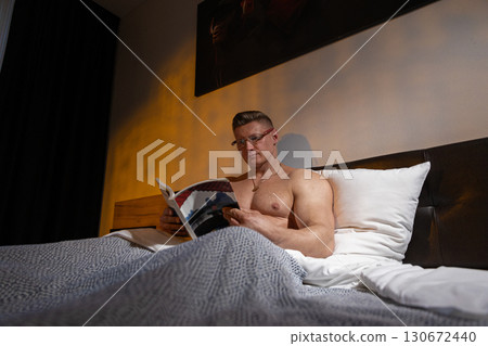 A shirtless man is comfortably sitting in bed wearing glasses and deeply focused on reading a book. The soft lighting creates a warm and inviting atmosphere. A shirtless man is comfortably sitting in bed wearing glasses and deeply focused on reading a book. The soft lighting creates a warm and inviting atmosphere. 130672440