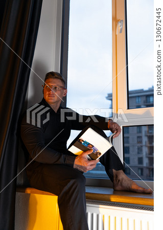 man in a black shirt and glasses sits barefoot on a windowsill deeply engrossed in a book. The backdrop shows a modern cityscape at dusk creating a serene atmosphere. 130672445