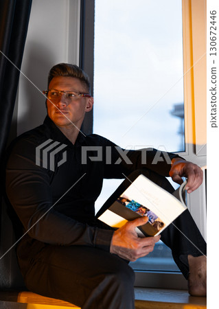 man dressed in a black shirt and glasses sits on a windowsill absorbed in reading a book. The warm glow of evening light adds a cozy atmosphere to his quiet moment. man dressed in a black shirt and glasses sits on a windowsill absorbed in reading a book. The warm glow of evening light adds a cozy atmosphere to his quiet moment. 130672446