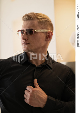 man wearing a black shirt and stylish glasses stands confidently showcasing his thoughtful expression. Soft warm lighting enhances the atmosphere creating a captivating indoor setting. 130672452