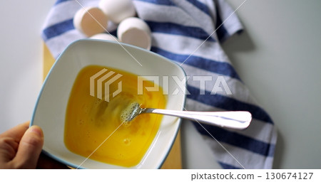 Closeup of hand whisking egg mixture in white bowl with fork, striped kitchen towel and egg shells nearby, top view breakfast preparation. Closeup of hand whisking egg mixture in white bowl with fork, striped kitchen towel and egg shells nearby, top view breakfast preparation. 130674127