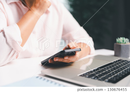 Close-up shot, woman sits at a wooden table using her smartphone, with her finger touching the phone screen as she navigates mobile banking, online shopping, and social media networks. Close-up shot, woman sits at a wooden table using her smartphone, with her finger touching the phone screen as she navigates mobile banking, online shopping, and social media networks. 130676455