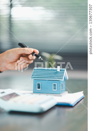 Real estate agent and customer are discussing contract to buy house, with house model in background symbolizing real estate, insurance, and loan arrangements involved in process. Real estate agent and customer are discussing contract to buy house, with house model in background symbolizing real estate, insurance, and loan arrangements involved in process. 130677307